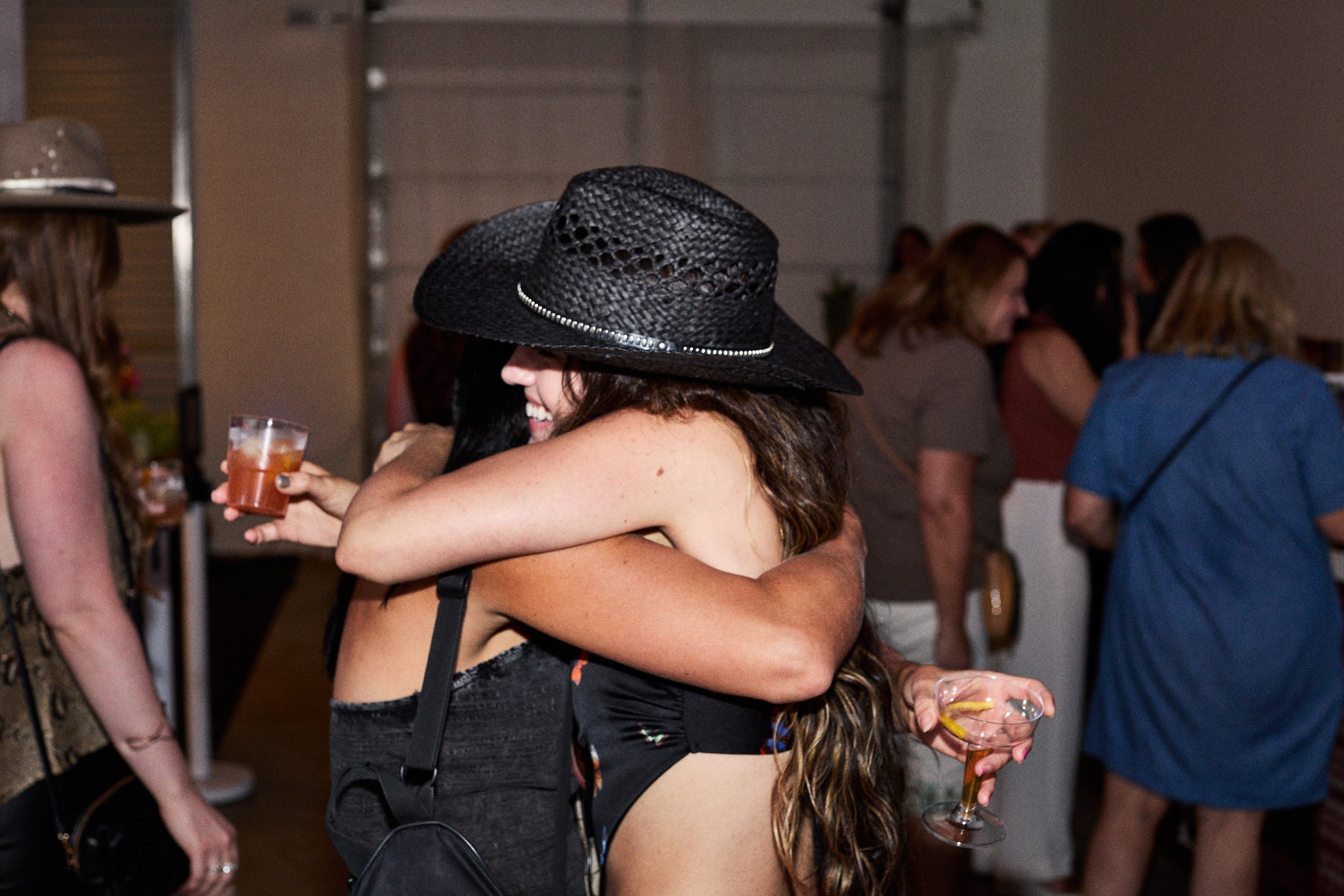 Two friends hugging at a party, holding non-alcoholic Curious Elixirs cocktails garnished with lemon peels.