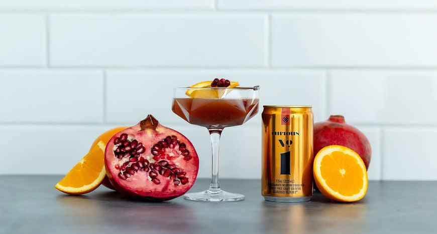 A photo of a small gold can of Curious Number 1 beside a glass containing the same drink. These are surrounded by halved pomegranates and oranges.