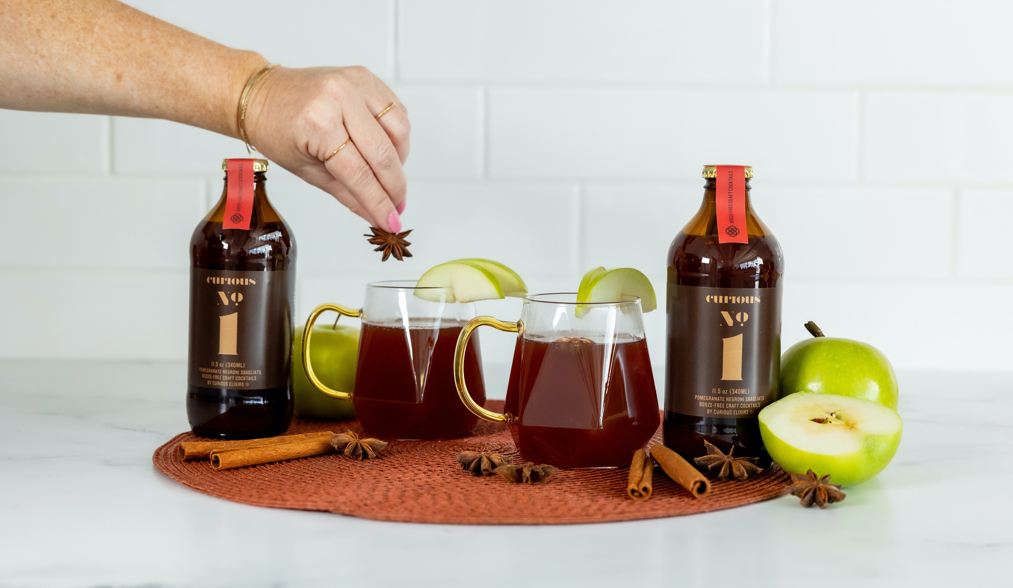 A photo of two brown glass bottles of Curious Number 1 beside two glass mugs full of a deep red drink. The drinks are garnished with green apple slices, and more apples and cinammon sticks rest around the beverages.