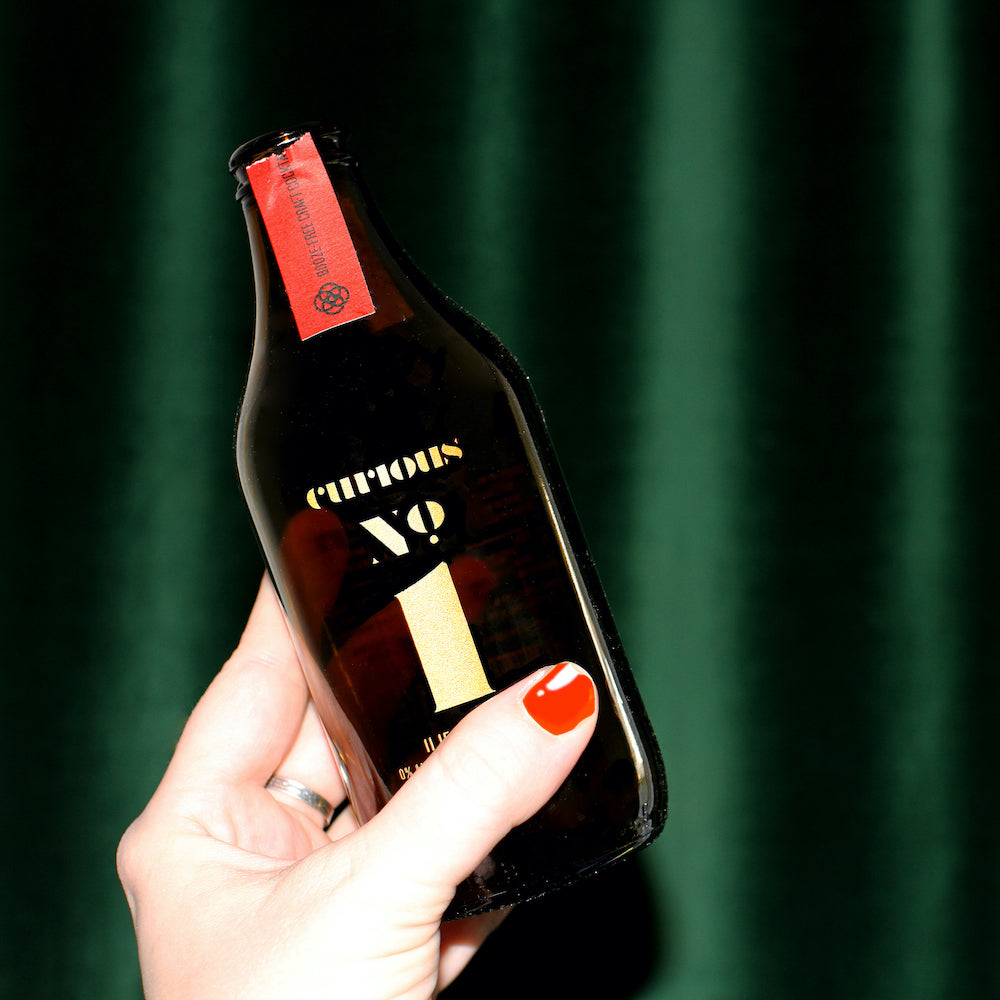An image of two brown glass bottles of Curious Number 1 on either side of a short glass full of a dark red cocktail. These all sit on a polished wood tabletop, with a fancy green and gold wall in the background.