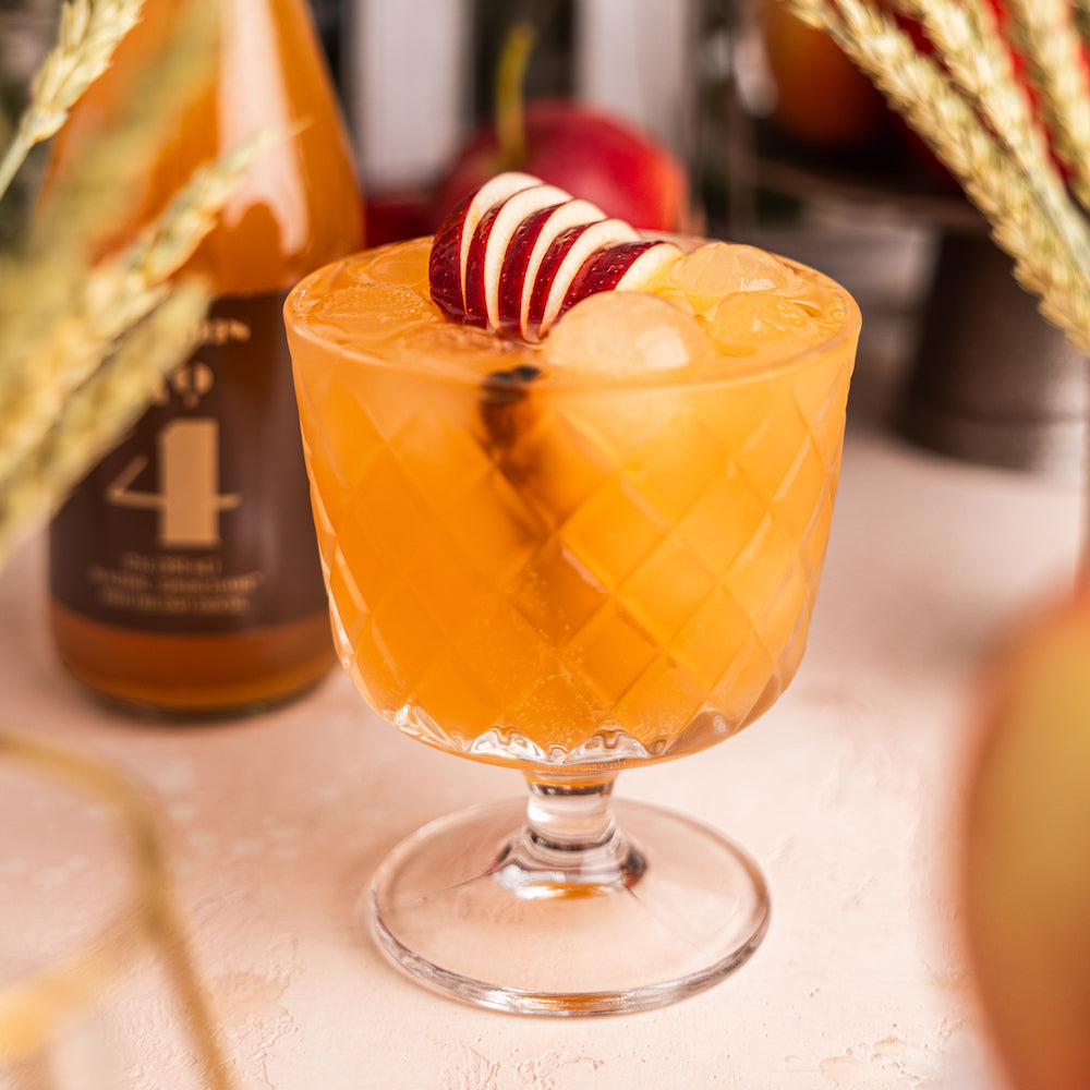 A photo of a glass goblet full of an orange drink garnished with apple slices and a cinnamon stick. In the background is a bottle of Curious Number 4.