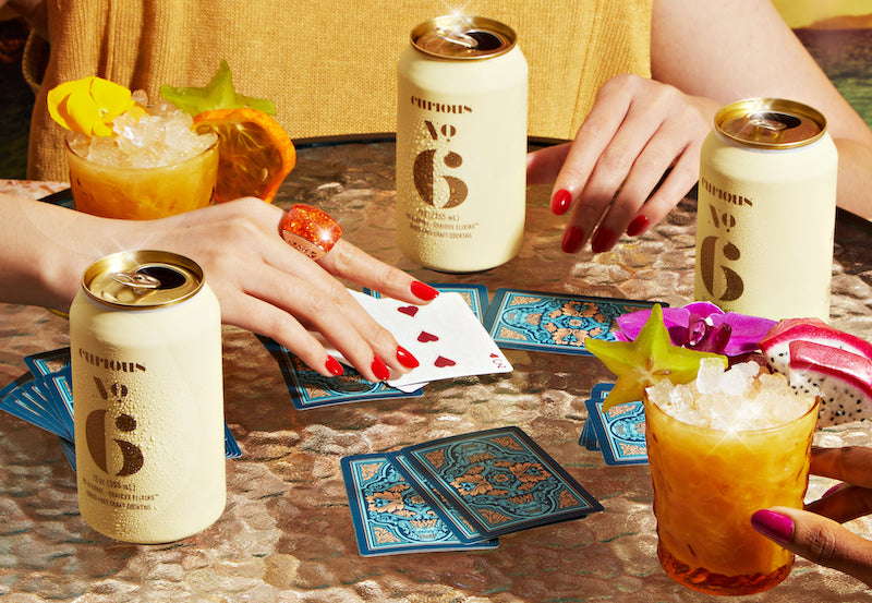 A photo of two people's hands as they play cards on a glass patio table. They are drinking from cream-colored cans of Curious Number 6. There are also glasses full of a creamy drink garnished with tropical fruits.