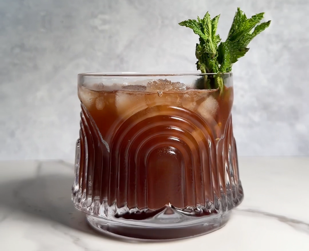 A short glass containing a reddish-brown drink served over ice and garnished with a sprig of mint.