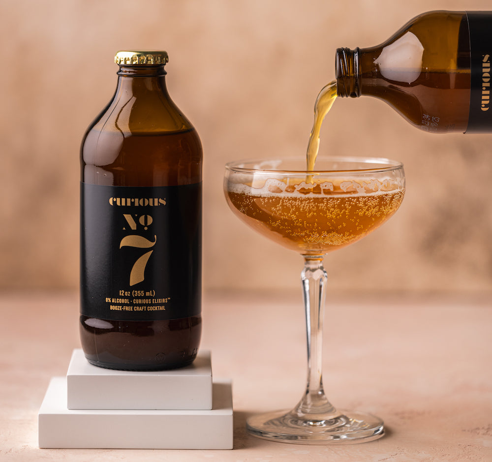 A photo of a brown glass bottle of Curious Number 7 sitting on small white blocks. To the right of the bottle is a coupe glass. Another bottle of Number 7 is being poured into the coupe glass. It looks similar to champagne.