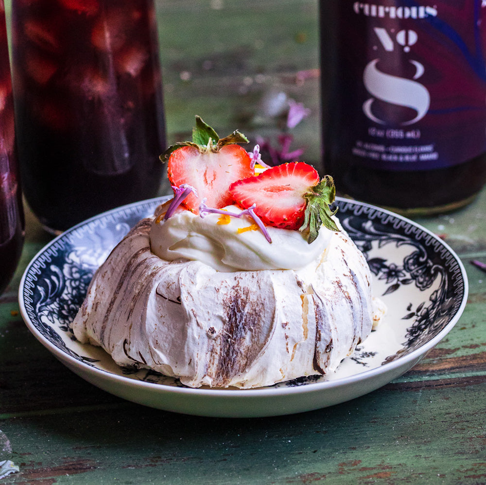 A photo of a pavlova served in a delicate dish and topped with sliced strawberries. In the background is a bottle of Curious Number 8, as well as a glass full of a deep burgundy drink.
