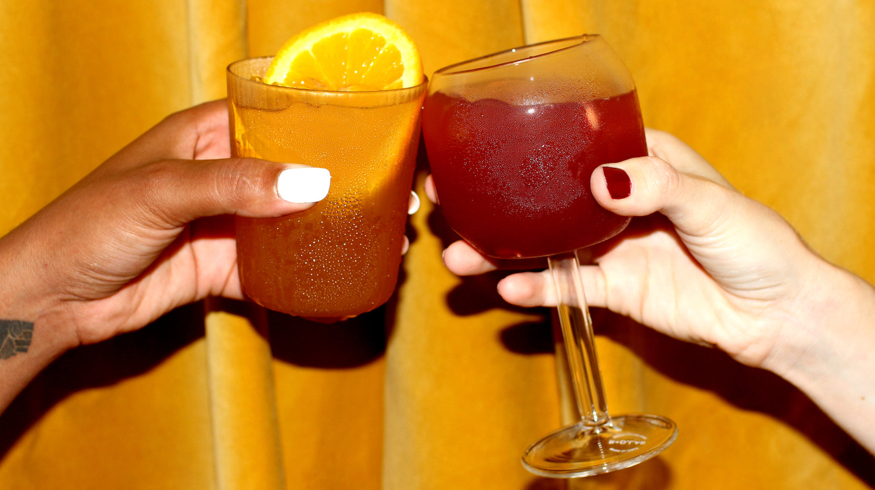 A photo of two people clinking their drinks together. One drink is dark orange and served in a short glass with an orange slice, while the other is a red beverage in a wine glass.