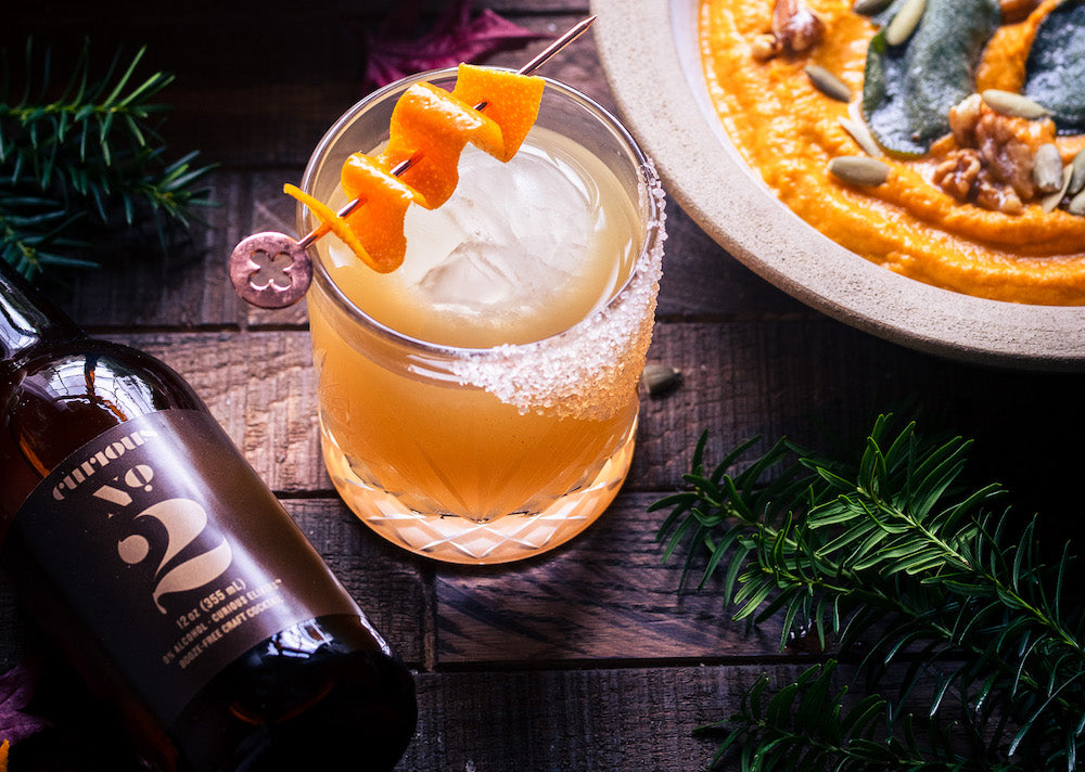 A photo of a glass on a wooden tabletop. It is filled with a pale orange beverage, served over a large ice cube and garnished with orange peel. To the left of the glass is a bottle of Curious Number 2. To the right is a bowl of pumpkin soup.