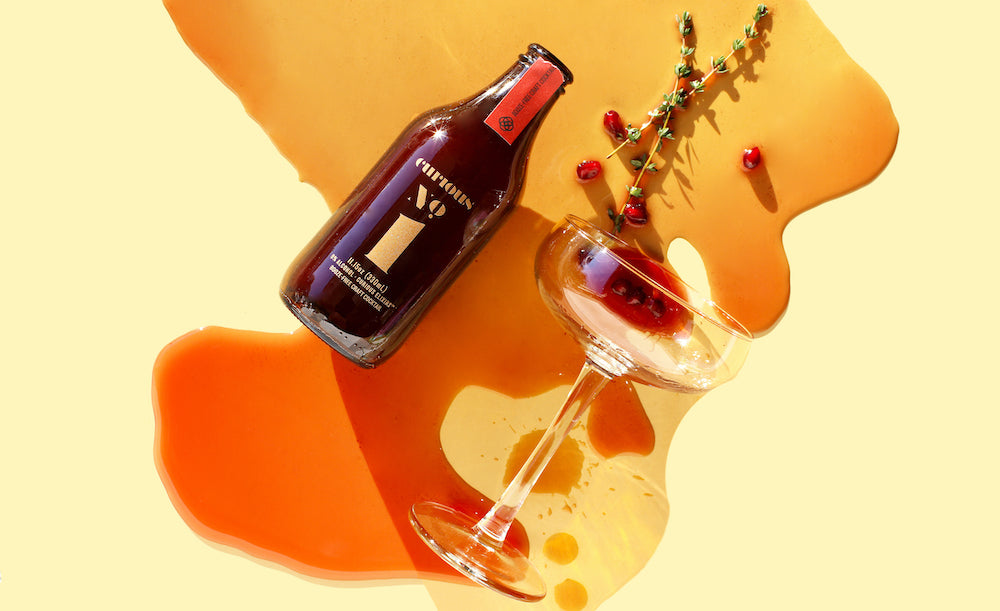 A photo looking down at a brown glass bottle of Curious Number 1 lying beside a coupe glass. The glass, also on its side, has spilled a dark red drink all over, but it still holds a sprig of fresh thyme and a few pomegranate seeds.
