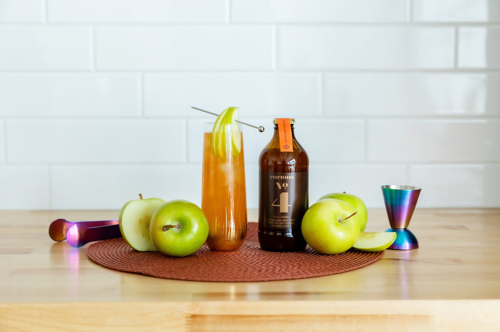 Curious No. 4 bottle on a table beside a garnished glass with apple slices on a skewer, surrounded by fresh green apples and cocktail mixing utensils.