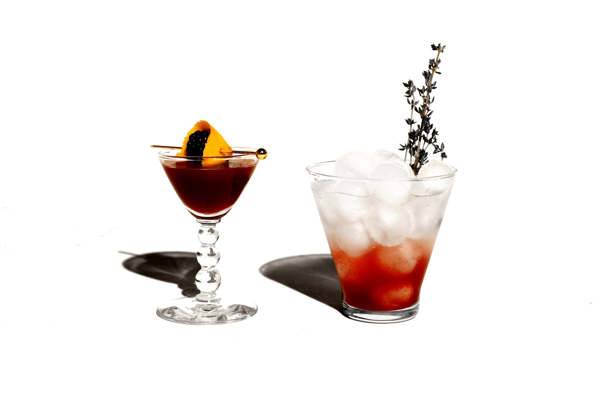 An image of a small port glass with a burgundy beverage in it, garnished with an orange peel and a blackberry. Beside this is a red spritz served over ice and garnished with a sprig of thyme.