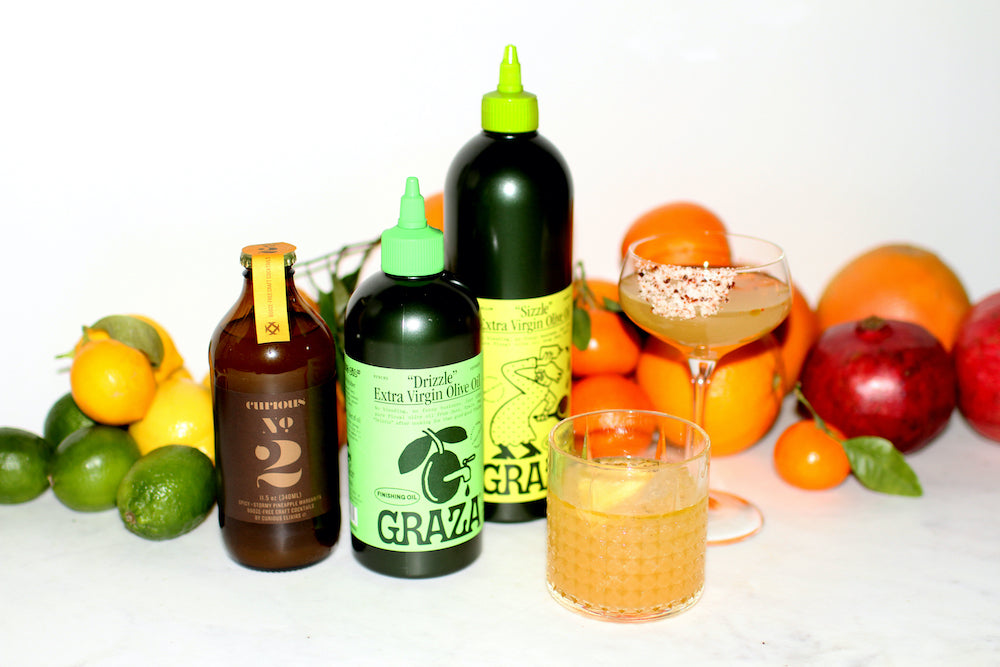 A photo of a brown glass bottle of Curious Number 2, two bottles of Graza olive oil, and two glasses with drinks that are pale orange in color, all surrounded by citrus fruits.
