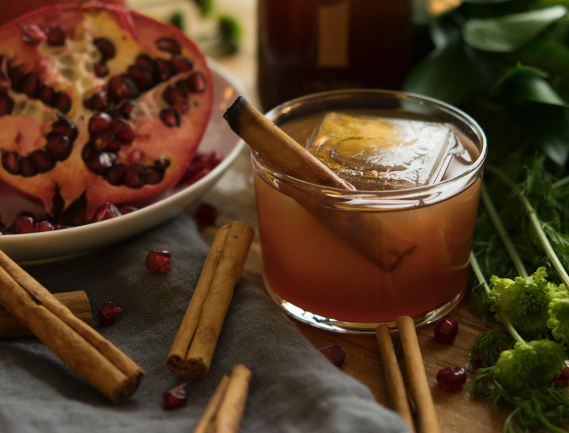 Beverage garnished with cinnamon and surrounded by pomegranates and herbs