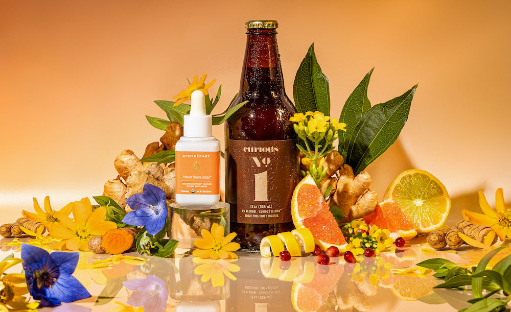 A photo of a bottle of Curious Number 1 beside a bottle of Apothekary bitters. These bottles are surrounded by a pile of fruit and flowers. The background is peach-colored.