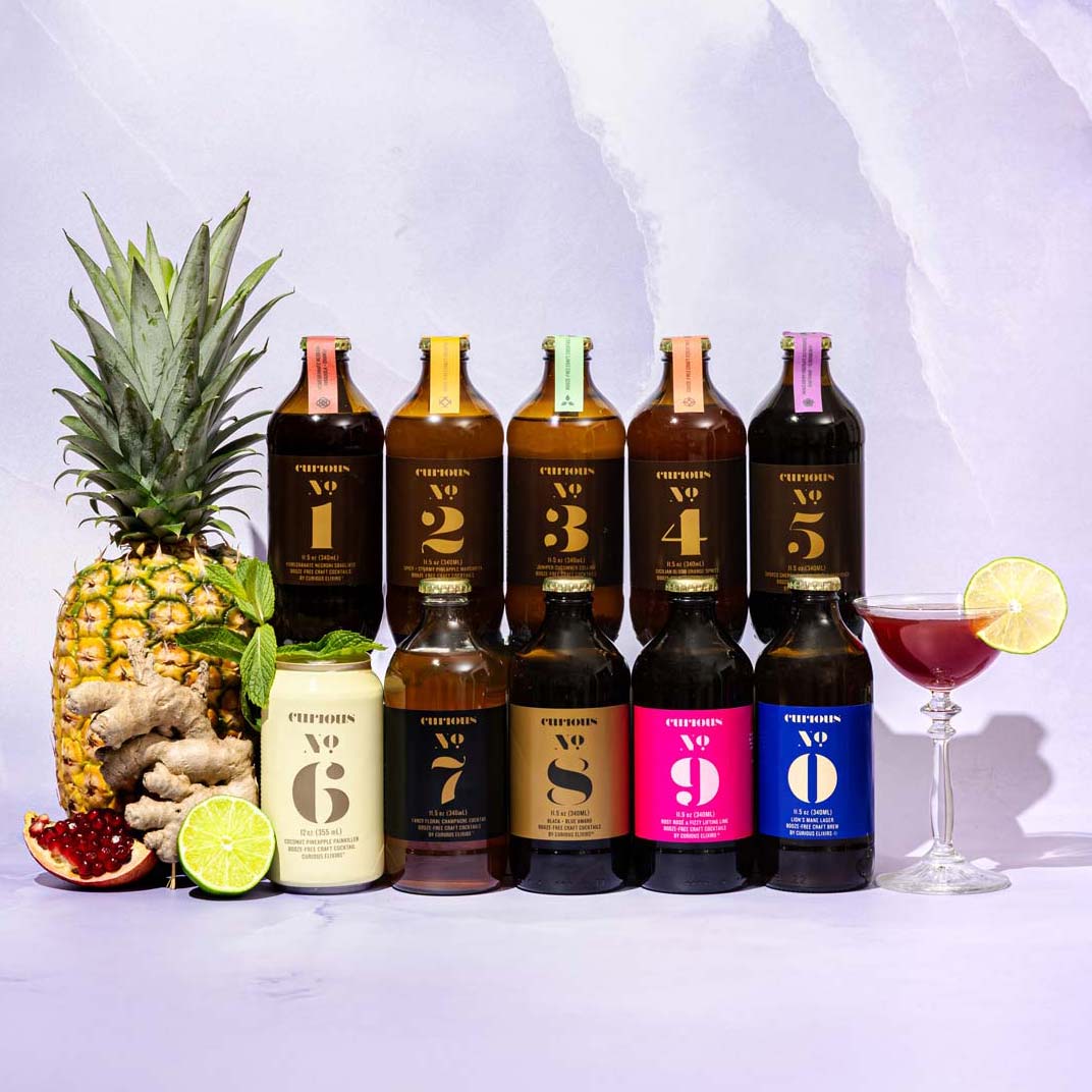 Set of all 10 Curious Elixirs bottles in two rows of 5, each brown bottle having a signature colorful label. Sitting to left and right are a pineapple, ginger, and a burgundy-colored nonalcoholic cocktail on a light marble background