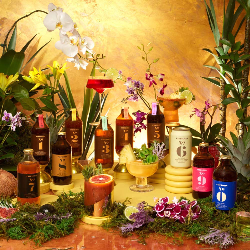 10 Nonalcoholic bottles of Curious Elixirs with colorful labels in a whimsical tableau of a decorative orchids, lilies, various plants, and moss.