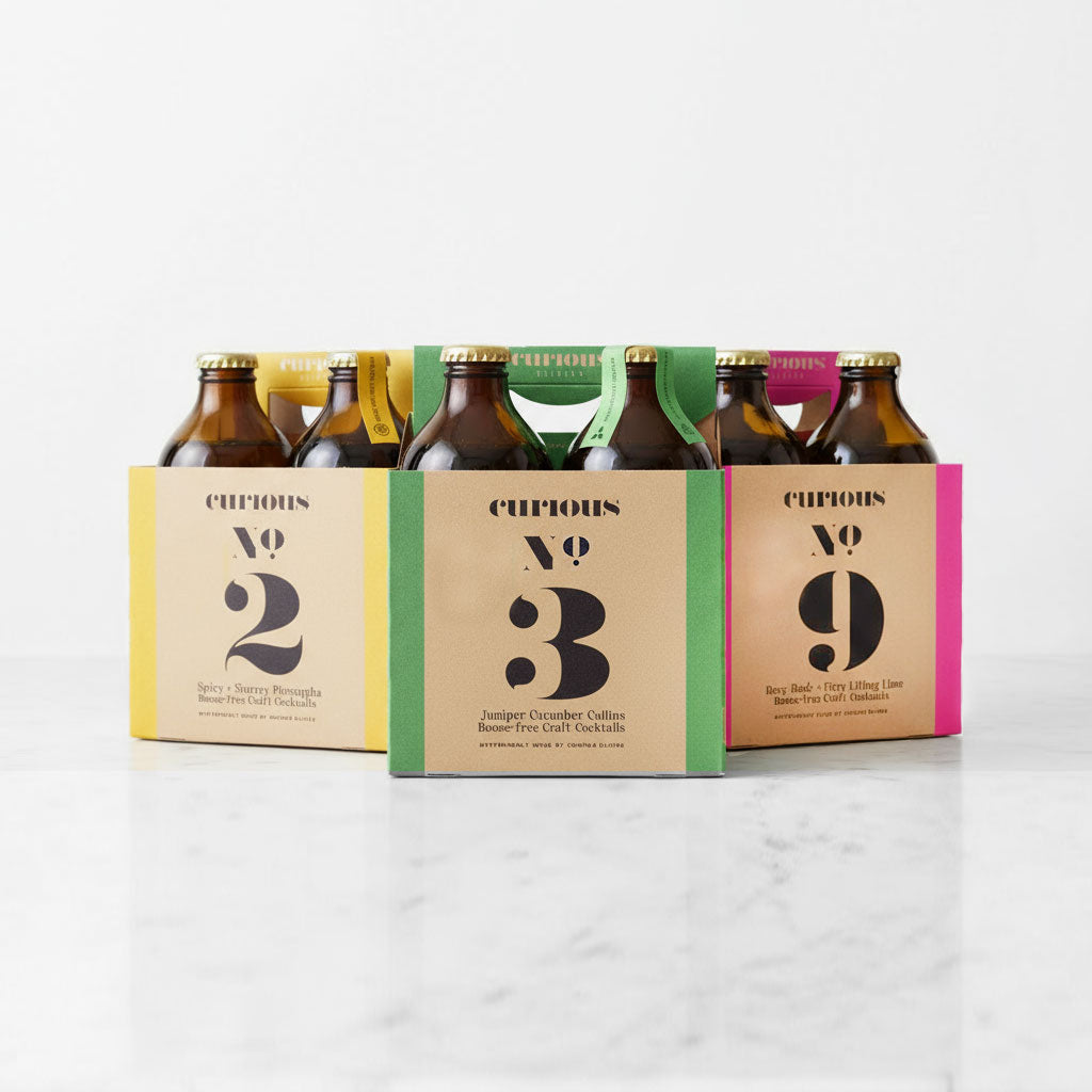 Three cardboard boxes with 4 bottles each of different flavors of Curious Elixirs sitting on a marble surface. The box on the left reads "Curious No. 2," the middle box "Curious No. 3 Juniper Cucumber Collins" and the right box "Curious No. 9"