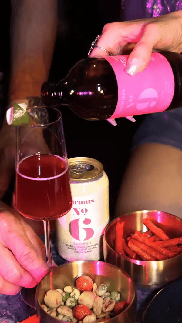 A hand with beautiful shimmery nail polish pours a bright pink bottle of Curious Elixir No. 9 into a champagne flute with a twist of lime peel on the rim. A can of Curious Elixir No. 6 rests on the table with bar snacks visible.